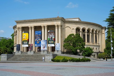 A magnificent theatre building with posters on pillars and trees in the foreground, Lado