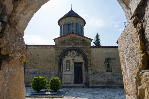 Through a stone arch, you look at an old church entrance and green courtyard, Motsameta monastery,