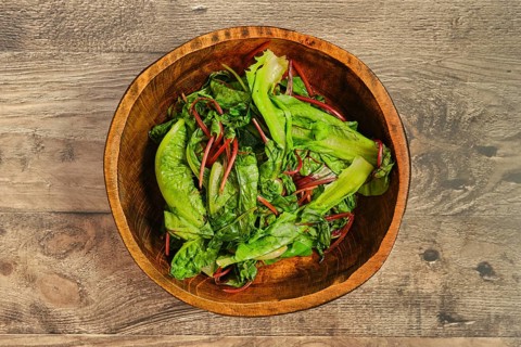 A wooden bowl filled with steamed greens on a rustic wooden tabletop. The leafy vegetables,