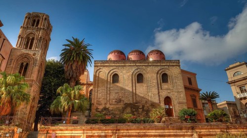 Side view of an Arab-Norman building with red domes and palm trees in sunny weather, Palermo, San