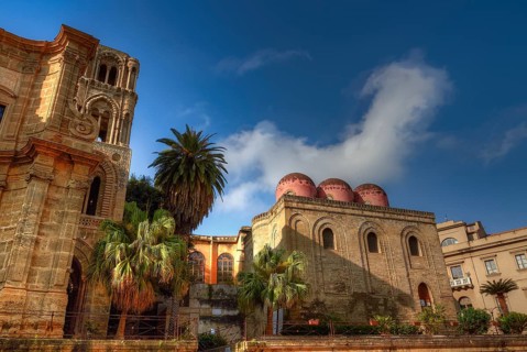 Arab-Norman historic building with red domes surrounded by palm trees under clear blue sky,