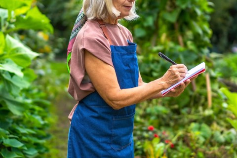 Senior woman in apron and colorful scarf jotting notes in a notepad with a pen while standing in a