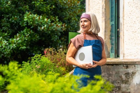 Senior woman in apron and headband holding a laptop outdoors in her garden, smiling and looking