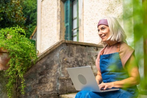 Senior woman enjoying remote work from a peaceful garden setting, smiling while typing on a laptop,