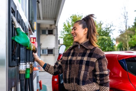 Woman smiling at a gas station while holding a fuel pump nozzle beside her red car in a plaid