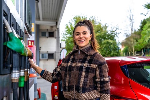Smiling young woman in a plaid jacket and protective glove refuels her red car at an urban gas