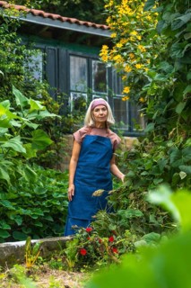 Senior woman in an apron and headband standing in a lush home vegetable garden, smiling while