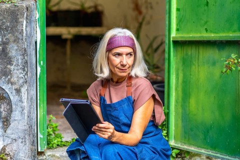 Senior woman artist in an apron and headband working on a digital tablet outside a rustic green