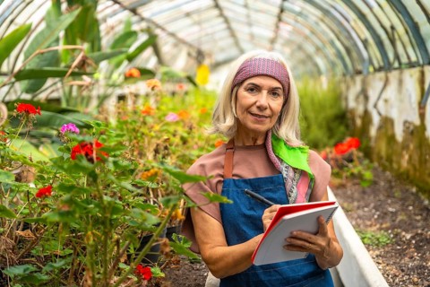 Senior woman wearing an apron and headband smiling, standing inside a botanical greenhouse while