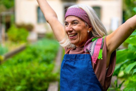 Senior woman in a bandana and apron raises her arms in a sunlit garden, smiling with joy and