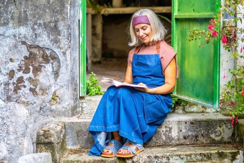 Senior woman in denim apron sits on old stone steps in a sunny garden, peacefully reading a book,