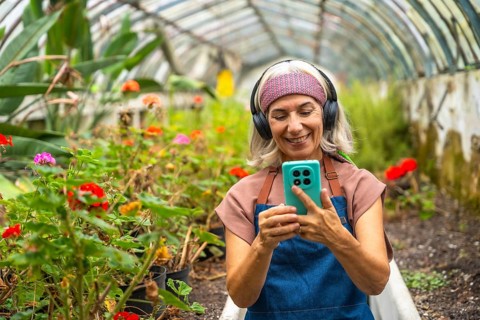Senior woman wearing headphones and apron, smiling while looking at her smartphone and interacting