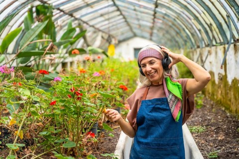 Senior woman smiling, wearing an apron and headphones, happily tending to potted plants and flowers