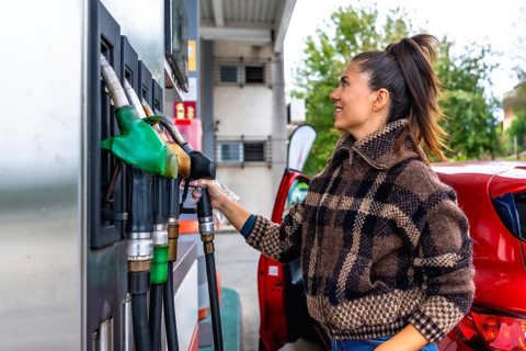 Woman in a casual plaid jacket holding a fuel pump nozzle, refueling her red car at a gas station,