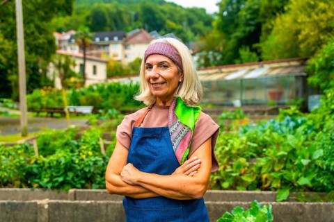 Senior woman wearing an apron and bandana standing with arms crossed in a vibrant community garden,