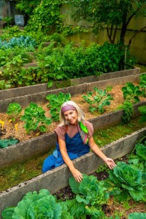 Senior woman in overalls smiling while tending raised bed allotment, harvesting organic vegetables