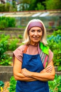 Senior woman wearing an apron and headscarf, standing with arms crossed in a lush community