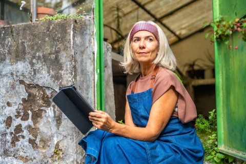 Senior woman in apron sitting by a greenhouse entrance, holding a digital tablet and looking