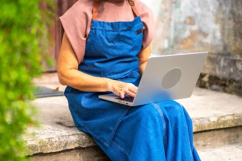 Senior woman in a blue apron working on a laptop outdoors, managing her small business and staying