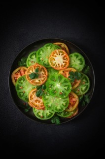 Sliced green tomatoes, on a dark surface, garnished with herbs and salt, top view
