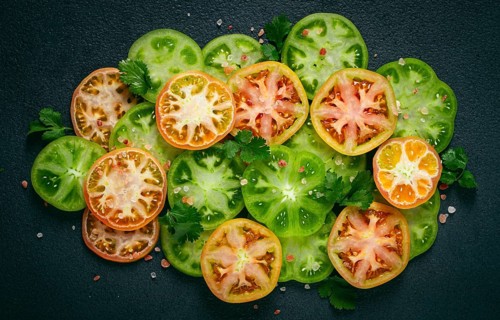 Sliced green tomatoes, on a dark surface, garnished with herbs and salt, top view