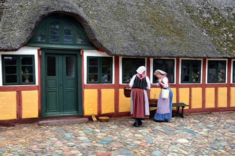Two women in older farm costumes in front of a half-timbered house with a thatched roof and on