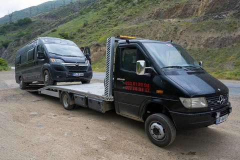 Tow truck transporting van on a rural road with green hills in the background, camper being towed,