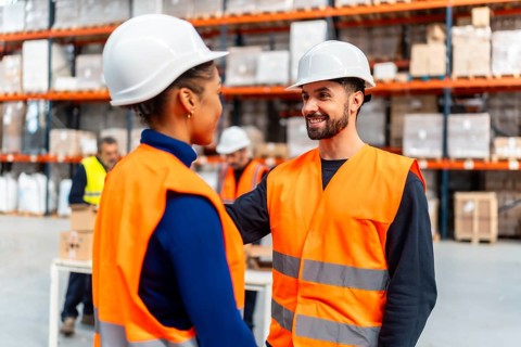 Two diverse warehouse workers wearing safety hard hats and vests are confidently smiling and