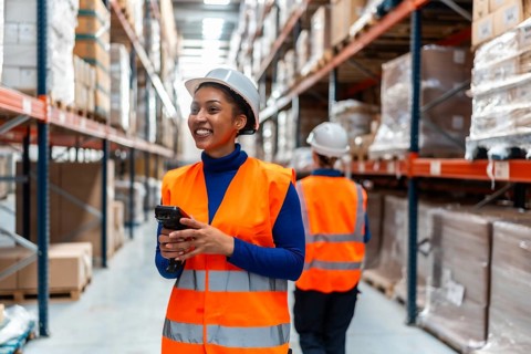 Young woman worker smiling, wearing a safety vest and hardhat, holding a barcode scanner while