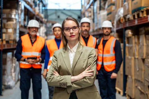 Confident female manager with arms crossed leading a diverse warehouse logistics team in a busy