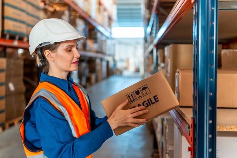 Woman warehouse worker wearing safety helmet and vest, inspecting a cardboard box with fragile