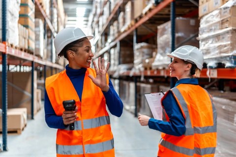 Two smiling female logistics workers wearing hard hats and safety vests collaborating while working