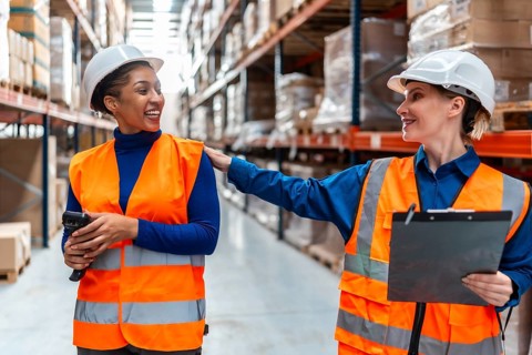 Two diverse women warehouse workers in safety vests and hard hats smiling and collaborating,