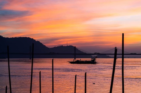 A boat sails on the river Brahmaputra during sunset, with the silhouette of a cable-stayed bridge