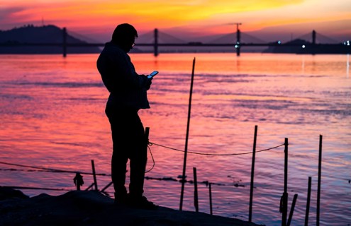 A person browsing his mobile phone by the Brahmaputra riverside, during sunset, on November 4, 2025