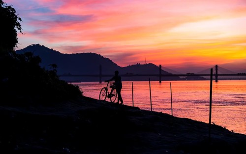 A man pushes his bicycle along the Brahmaputra riverbank during a vibrant sunset, with the
