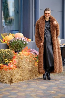 A woman stands next to the autumn installation, wearing a brown coat and black dress. She poses