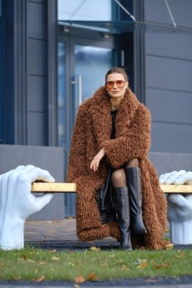 A stylish woman wearing a brown fur coat and black knee-high boots poses on a unique bronze bench