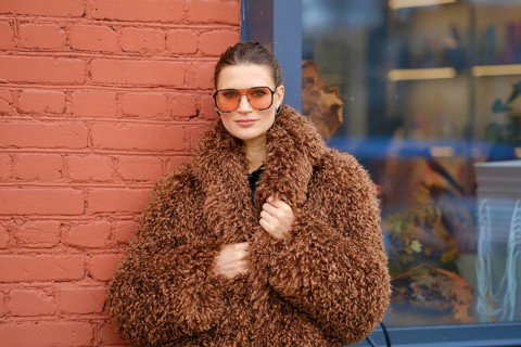 A woman poses confidently in a warm, brown, fluffy coat and oversized sunglasses. She stands