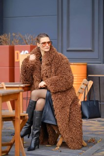 A stylish woman is seated at a wooden table outside a cafe, wearing a fluffy brown coat and