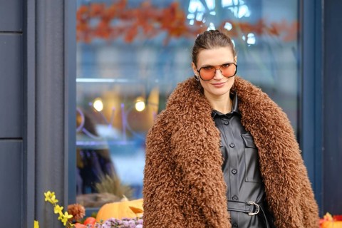 A woman wearing a brown furry coat and sunglasses stands outside a cozy cafe filled with autumn