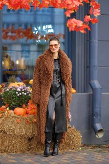 A woman dressed in a stylish brown fuzzy coat and black leather dress stands next to a seasonal