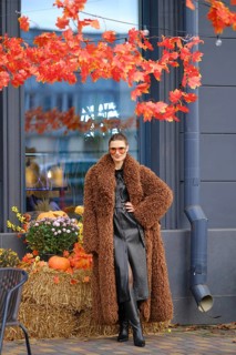 A stylish model stands near a store with bright autumn leaves overhead. She wears a brown fur coat