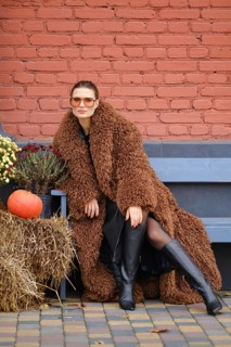 A woman wearing a brown fuzzy coat and knee-high black boots poses confidently outdoors. She is