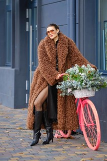A fashionable woman stands against a modern building, wearing a cozy brown coat and tall black