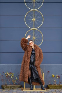 A woman stands confidently near a striking bike wheel art piece. She wears a fluffy brown coat and