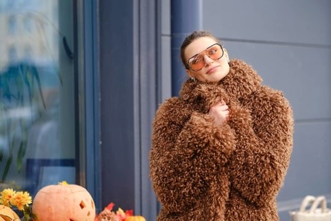 A young woman stands outside a shop, dressed in a fluffy brown coat and stylish sunglasses.