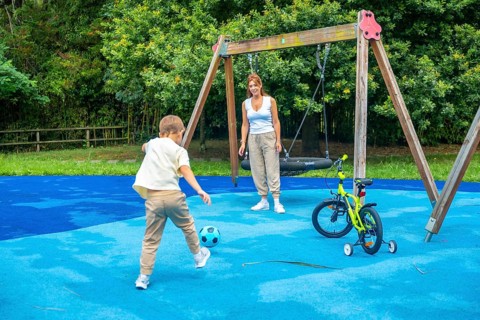 Young boy kicking a soccer ball on a playground while mother watches, with a swing set and bicycle