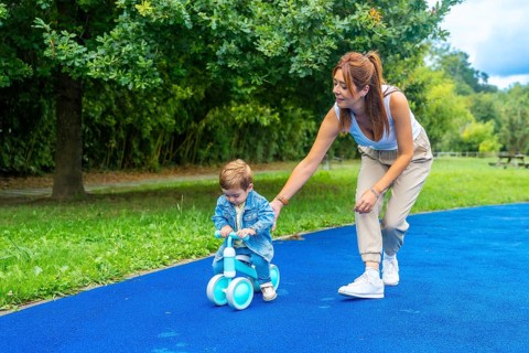 Young mother helping her son learn to ride a balance bike on a blue surface in a green park,