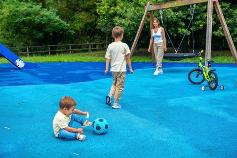 Two brothers are playing with a ball and a bicycle at the playground while their mother watches